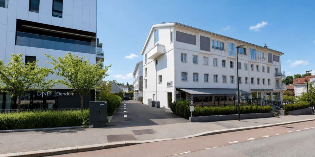 Moderne hvit bygning med store vinduer og grønt område foran, i bymiljø.