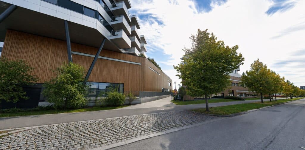 Moderne klinikkbygning med tre og glass, omgitt av trær og en gangvei i et bymiljø.