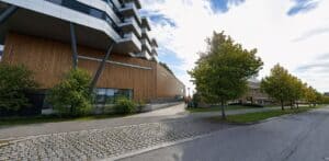 Moderne klinikkbygning med tre og glass, omgitt av trær og en gangvei i et bymiljø.