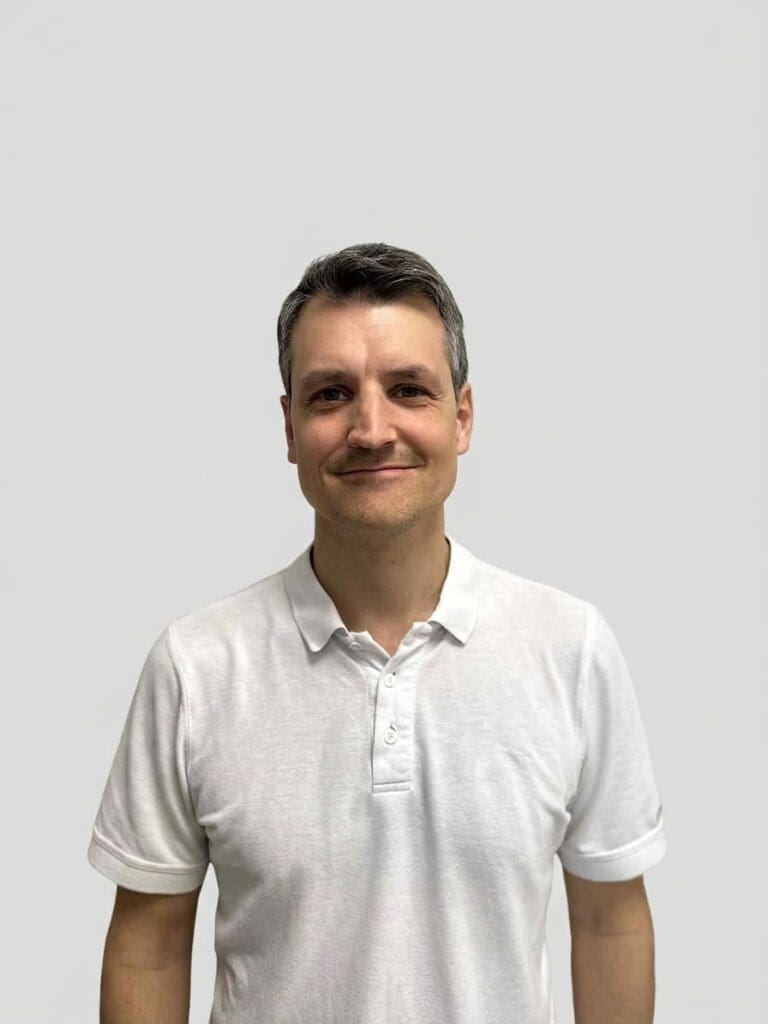 Mann i hvit polo-shirt smiler mot kamera mot en lys bakgrunn.