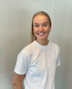Kvinne smilende i hvit t-skjorte foran lys vegg.