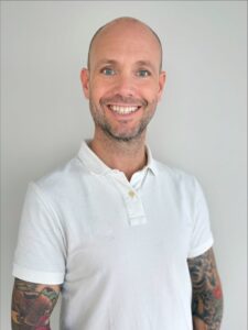 Mann med tatoveringer i hvit polo smiler mot kamera.