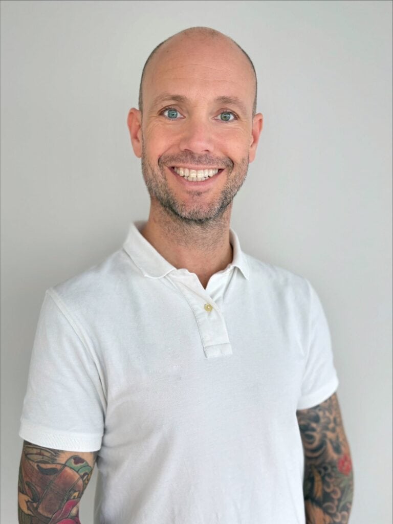 Mann med tatoveringer i hvit polo smiler mot kamera.