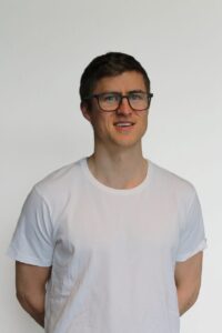 Mann med briller i hvit t-skjorte smilende mot kamera.