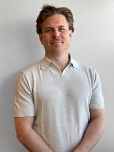 Mann smilende i hvit polo foran lys grå vegg.