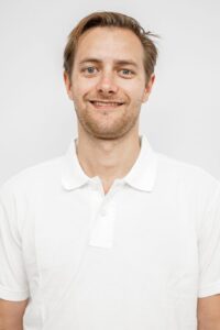 Mann med hvit polo-shirt smilende foran lys bakgrunn.