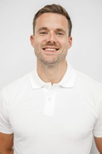 Mann med hvit polo smilende foran lys bakgrunn.