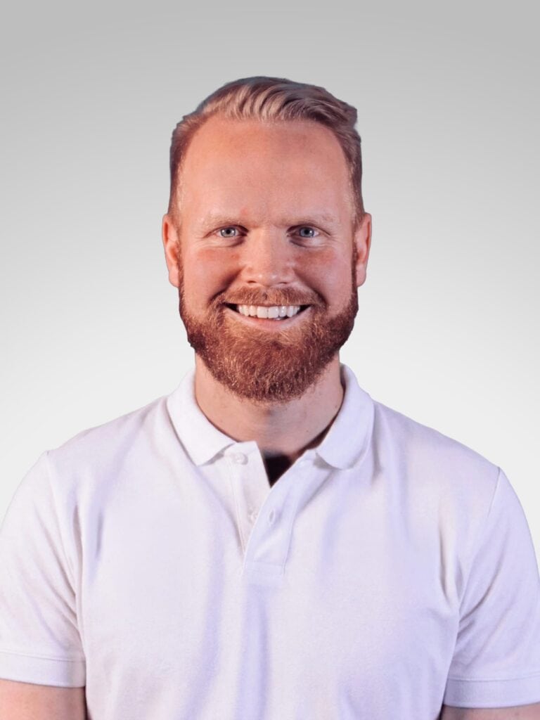 Mann med lyst hår og skjegg smilende i hvit polo, mot lys grå bakgrunn.