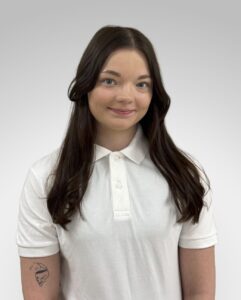 Ung kvinne med langt brunt hår i hvit polo-shirt, smilende mot kamera.