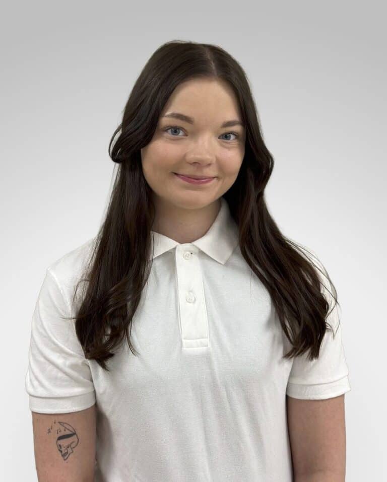 Ung kvinne med langt brunt hår i hvit polo-shirt, smilende mot kamera.