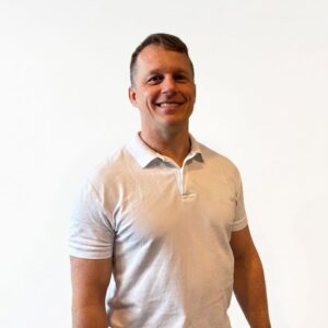 Mann i hvit polo-shirt smiler mot kamera mot en lys bakgrunn.