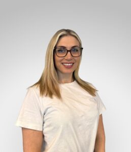 Kvinne med briller smilende mot kamera, hvit t-skjorte, mot lys grå bakgrunn.