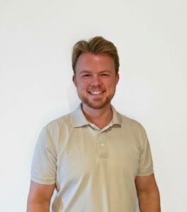Mann med lyst hår og beige polo shirt smilende mot hvit bakgrunn.