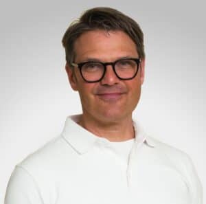Mann med briller i hvit skjorte smilende mot kamera.