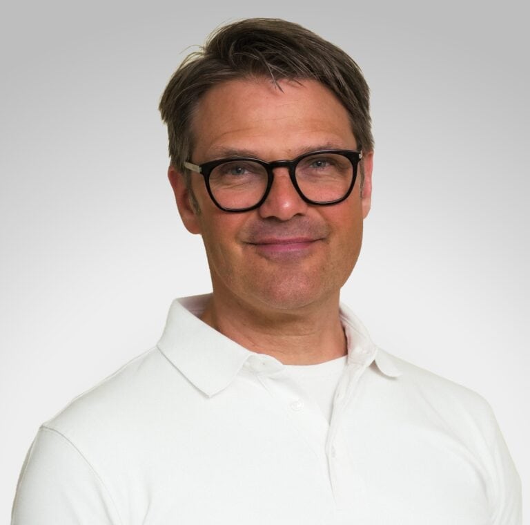 Mann med briller i hvit skjorte smilende mot kamera.