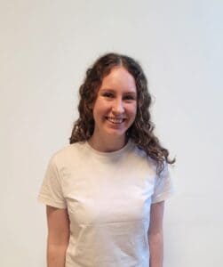 Smilende kvinne med krøllete hår i hvit t-skjorte mot lys bakgrunn.