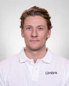 Kvinne i hvit Limba-uniform med blå øyne og lyst hår, smilende mot kameraet.