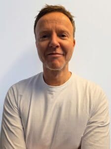 Mann med kort hår og hvit t-skjorte smilende mot kamera.