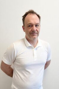 Mann i hvit polo shirt står mot hvit vegg, ser direkte inn i kameraet.