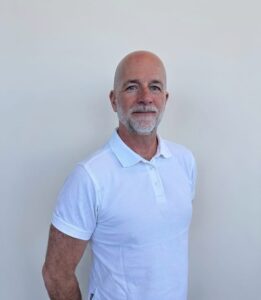Mann med hvit polo shirt smilende foran lys bakgrunn.