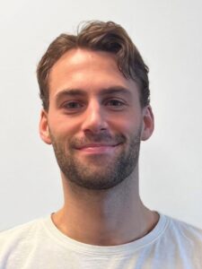 Liten portrett av Filip Angell, smilende mann med kort, brunt hår og skjegg, mot en lys bakgrunn.