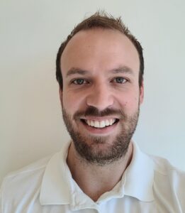 Mann med kort hår og skjegg smiler mot kamera, hvit bakgrunn.