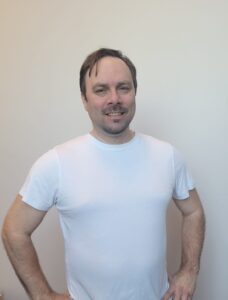 Mann i hvit t-skjorte smiler foran lys vegg.