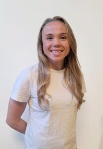 Smilende kvinne med langt, blond hår i hvit t-skjorte mot lys bakgrunn.