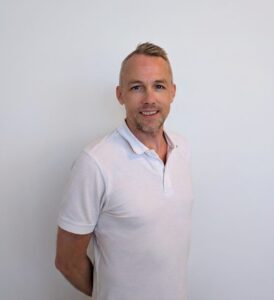 Kvinne smilende mann i hvit polo foran hvit vegg.