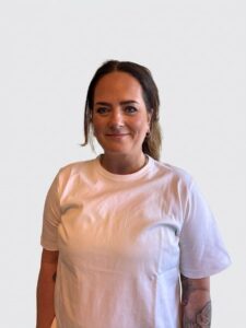 Kvinne med mørkt hår og hvit t-skjorte smiler mot kamera.