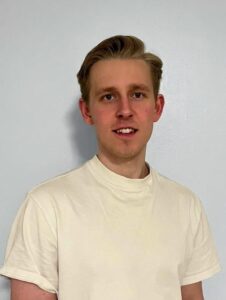 Mann i hvit t-skjorte smiler mot kamera, innendørs.