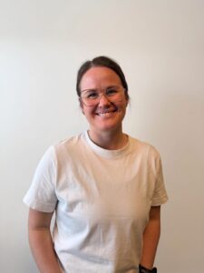 Kvinne med briller smilende mot kamera, hvit t-skjorte, innendørs.