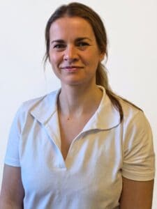Kvinne i hvit polo shirt smiler mot kamera, innendørs, helsepersonell.