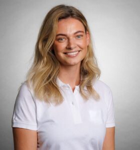 Smilende kvinne i hvit polo-shirt mot lys grå bakgrunn.