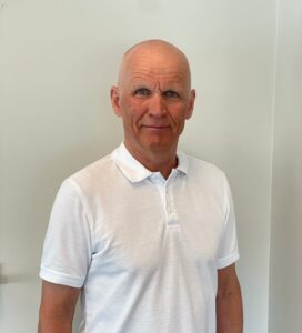 Kiropraktor smiler i klinikkmiljø, hvit polo, nærbilde.