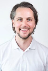 Mann med mørkt hår og skjegg smiler mot kamera, hvit bakgrunn.