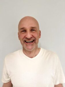 Smilende mann med hvit t-skjorte mot lys bakgrunn.