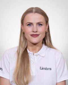 Kvinne i hvit Limbra-polo med lang, blond hår og gulløredobber, smilende mot kameraet.