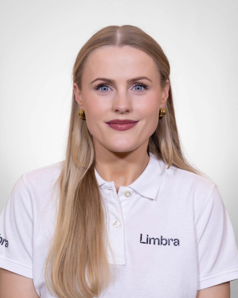 Kvinne i hvit Limbra-polo med lang, blond hår og gulløredobber, smilende mot kameraet.