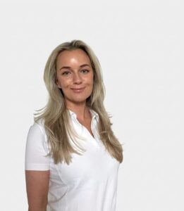 Kvinne med blond hår i hvit polo-shirt smilende mot kamera.