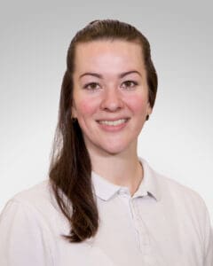 Kvinne smilende helsepersonell med hvit skjorte mot grå bakgrunn.