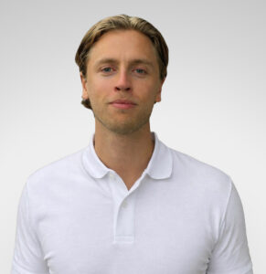 Mann med lyst hår og hvit polo, portrett mot lys grå bakgrunn.