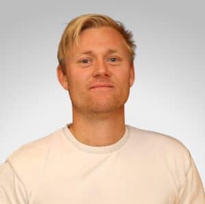 Mann med blond hår og hvit t-skjorte smilende mot kamera.