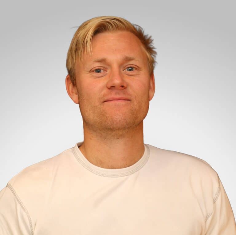 Mann med blond hår og hvit t-skjorte smilende mot kamera.