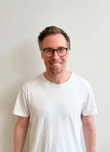 Mann med briller smilende i hvit t-skjorte mot lys bakgrunn.