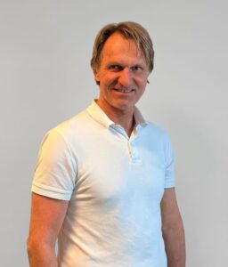 Kiropraktor smiler i hvit polo foran nøytral bakgrunn.