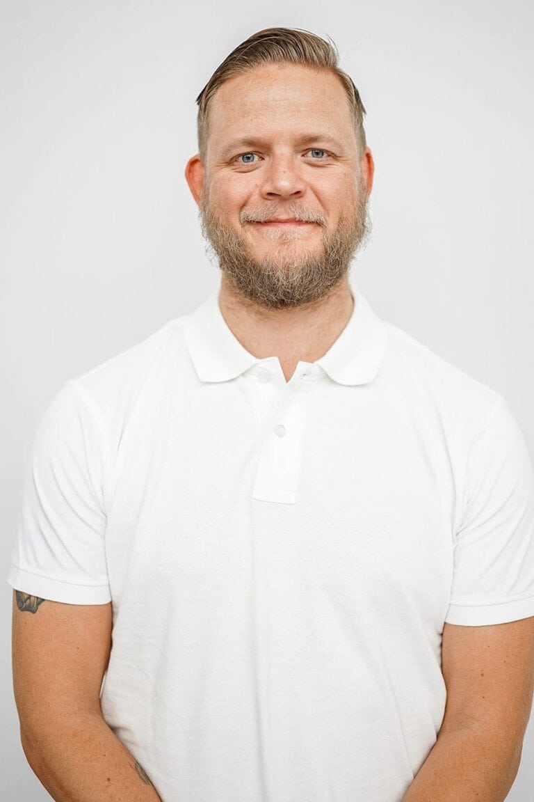 Mann med lyst hår og skjegg i hvit polo, smilende foran lys bakgrunn.