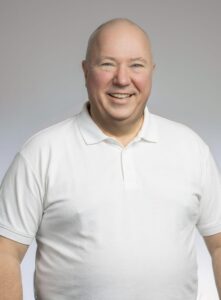 Mann med hvit polo smilende foran lys grå bakgrunn.