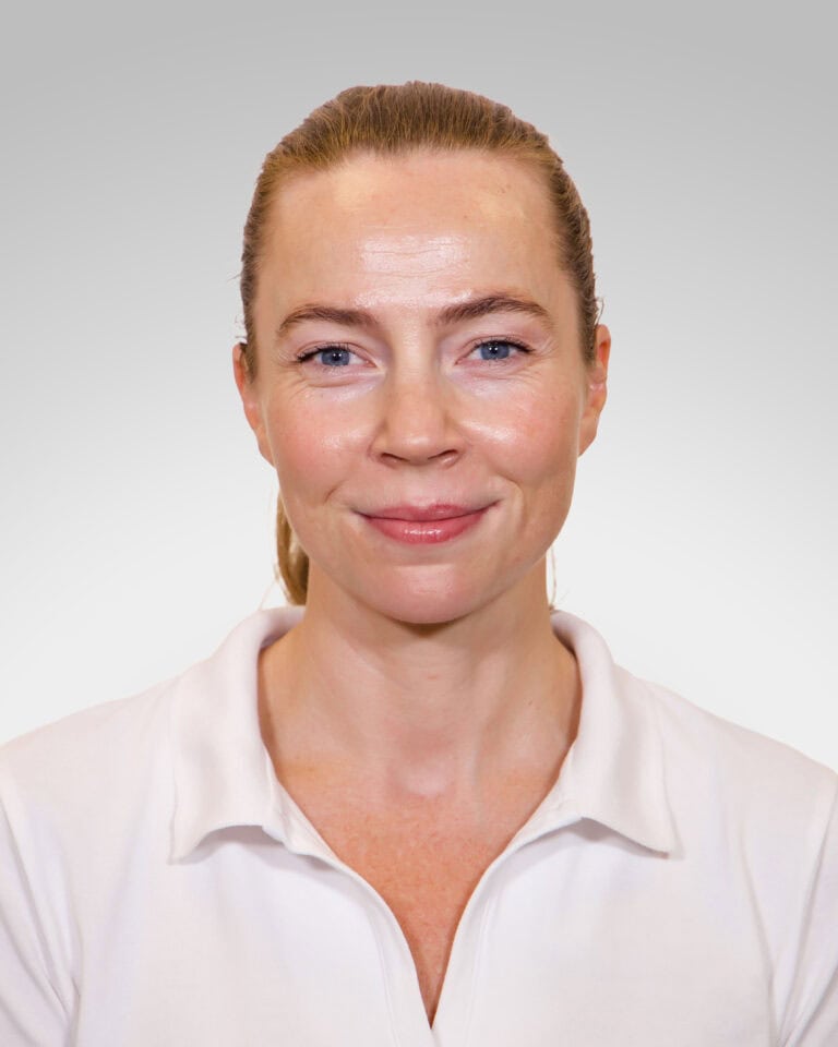 Kvinne med hvit skjorte smilende mot kamera, nøytral bakgrunn.