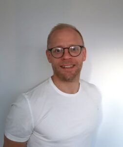 Mann med briller smilende i hvit t-skjorte foran lys bakgrunn.